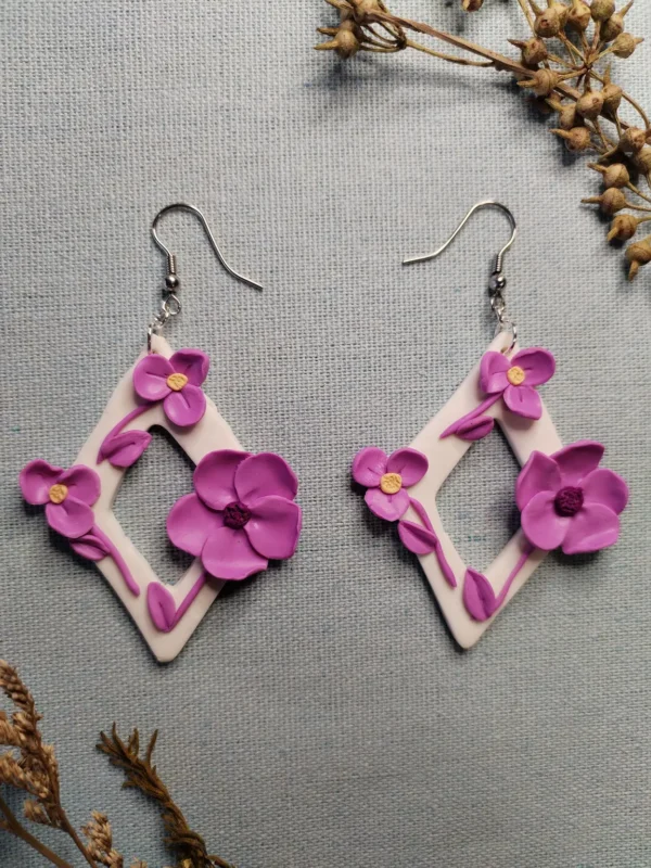 Handmade diamond-shaped polymer clay earrings with small purple flowers and purple leaves, displayed on a neutral background.