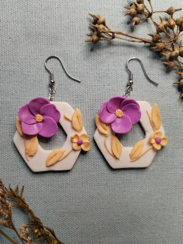 A pair of handmade hexagon-shaped polymer clay earrings featuring purple and peach flowers with leaves, displayed on a textured fabric background with dried botanical elements.