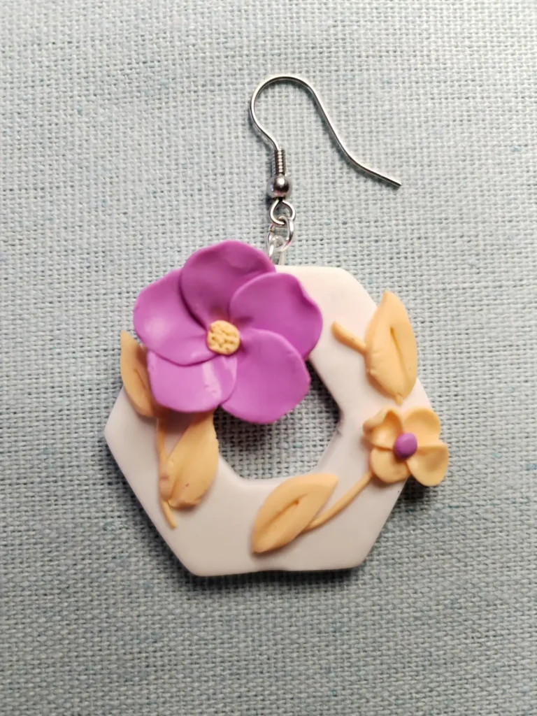 A pair of handmade hexagon-shaped polymer clay earrings featuring purple and peach flowers with leaves, displayed on a textured fabric background with dried botanical elements.