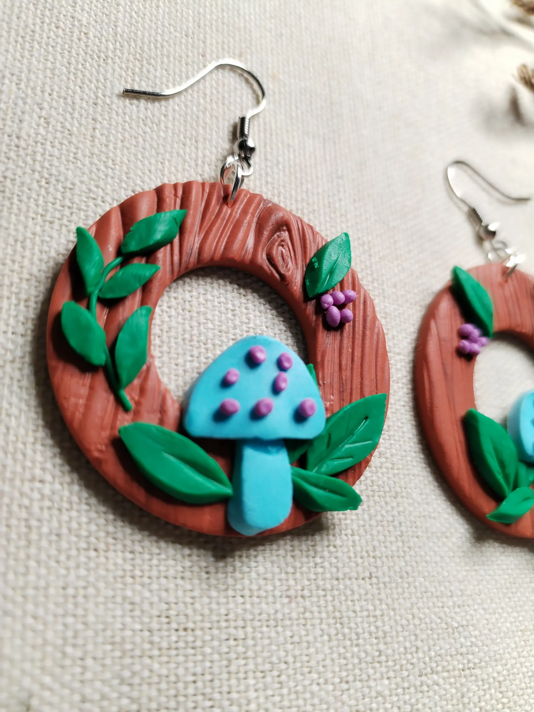 Handgemachte Ohrringe aus Polymer Clay mit türkisfarbenen Pilzen, grünen Blättern und lilafarbenen Beeren auf brauner Holzstruktur, inspiriert von einem Waldmotiv