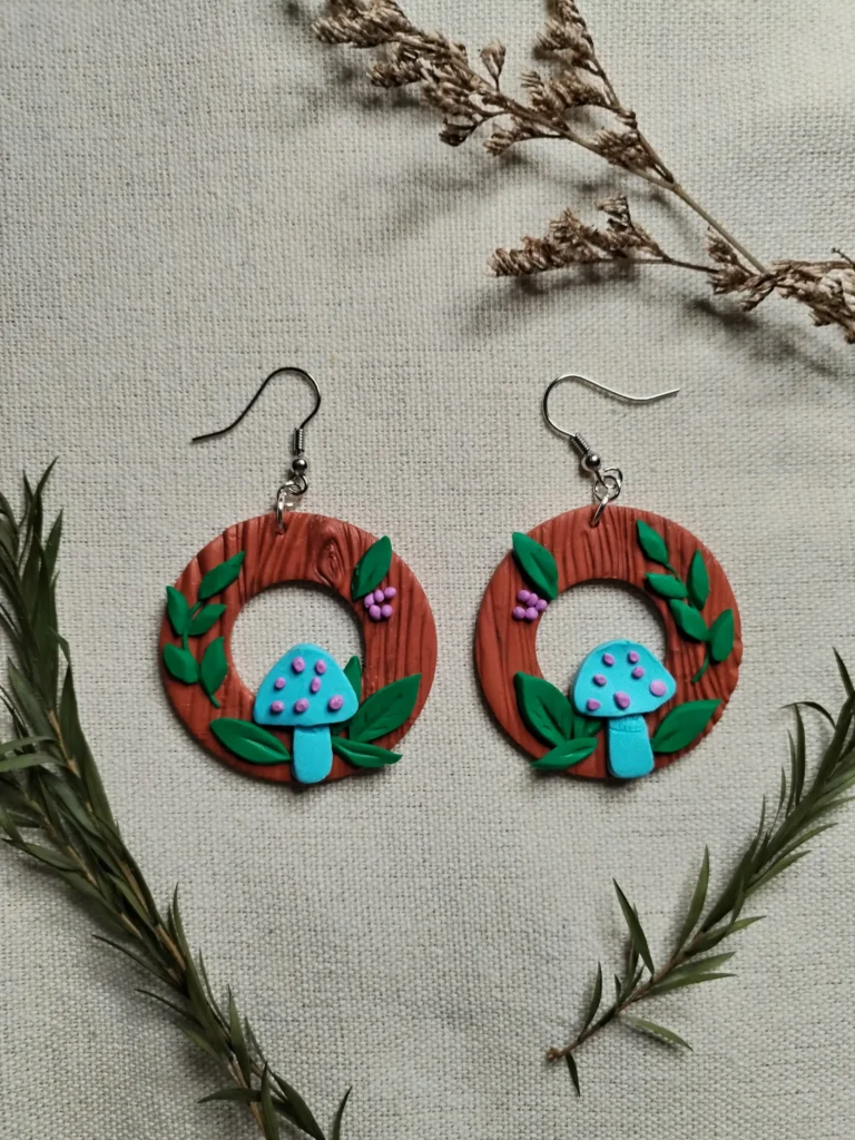 Handgemachte Ohrringe aus Polymer Clay mit türkisfarbenen Pilzen, grünen Blättern und lilafarbenen Beeren auf brauner Holzstruktur, inspiriert von einem Waldmotiv