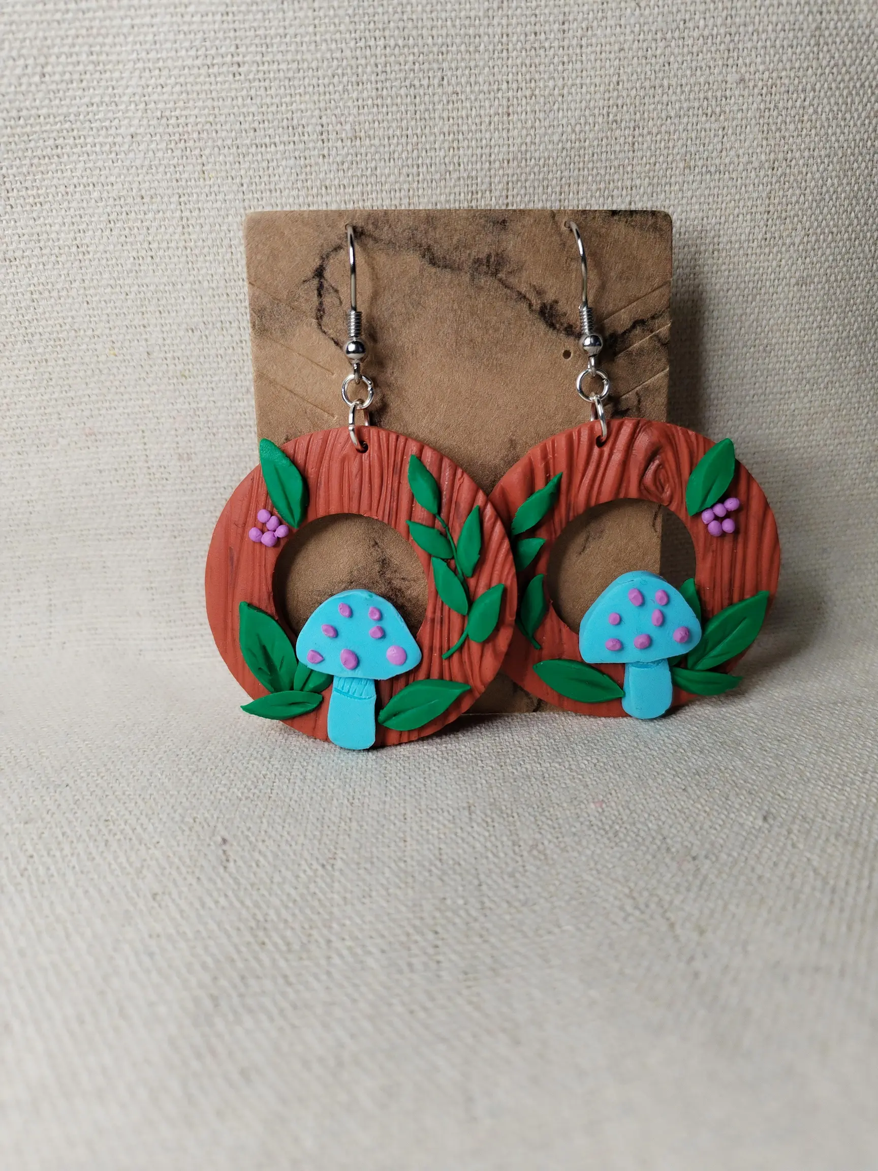 Handgemachte Ohrringe aus Polymer Clay mit türkisfarbenen Pilzen, grünen Blättern und lilafarbenen Beeren auf brauner Holzstruktur, inspiriert von einem Waldmotiv