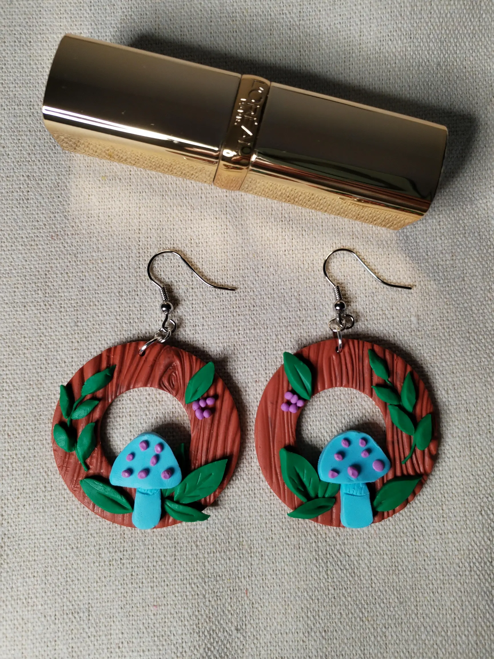 Handgemachte Ohrringe aus Polymer Clay mit türkisfarbenen Pilzen, grünen Blättern und lilafarbenen Beeren auf brauner Holzstruktur, inspiriert von einem Waldmotiv