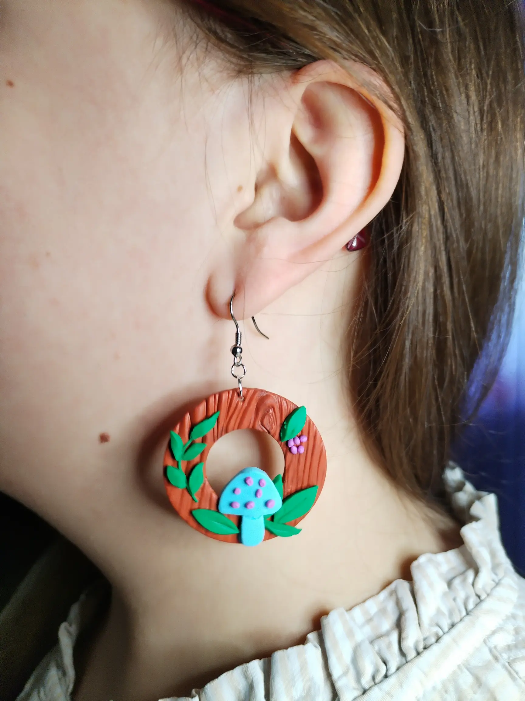Handgemachte Ohrringe aus Polymer Clay mit türkisfarbenen Pilzen, grünen Blättern und lilafarbenen Beeren auf brauner Holzstruktur, inspiriert von einem Waldmotiv